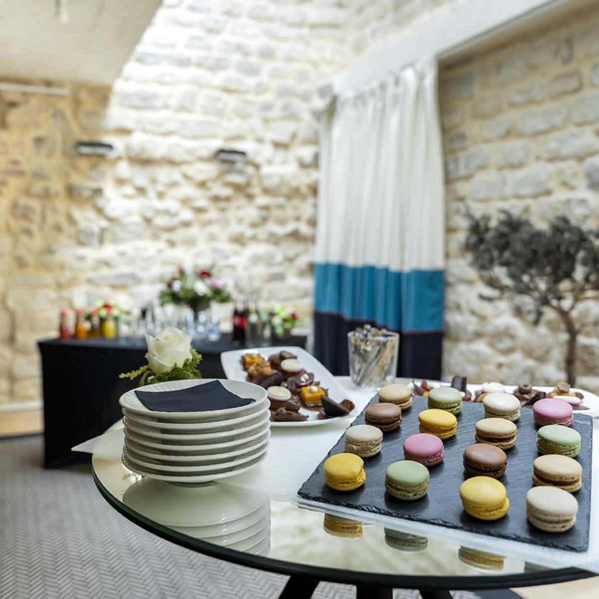 A table set with plates and colorful macarons, alongside a buffet featuring drinks and floral arrangements in a stone-walled room.