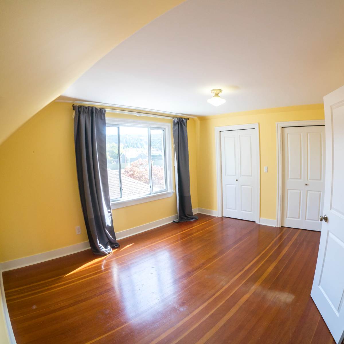 A yellow-walled room with wooden floors, two closed doors, and a window with gray curtains.