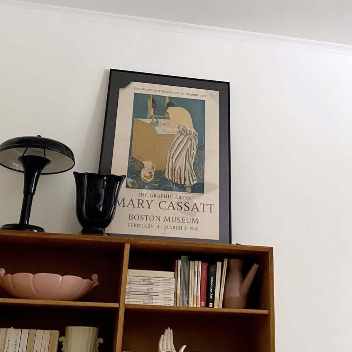 A framed poster of Mary Cassatt's graphic art hangs on a white wall above a wooden shelf filled with books and decorative items.