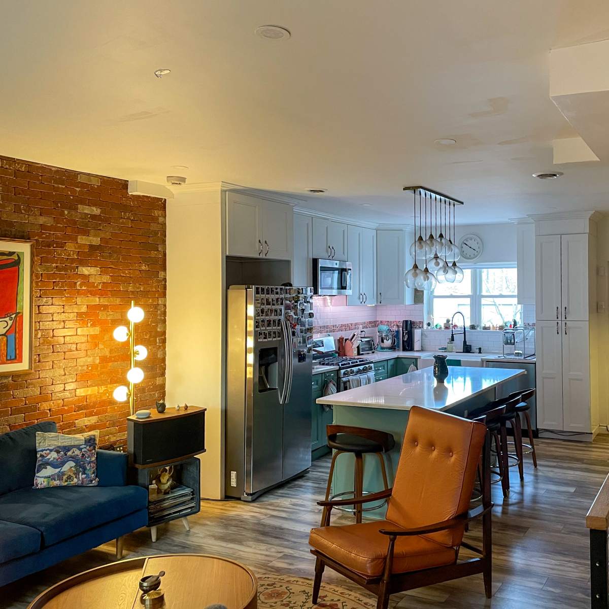 A modern open-concept kitchen and living area with a staircase on the right and a brick accent wall.