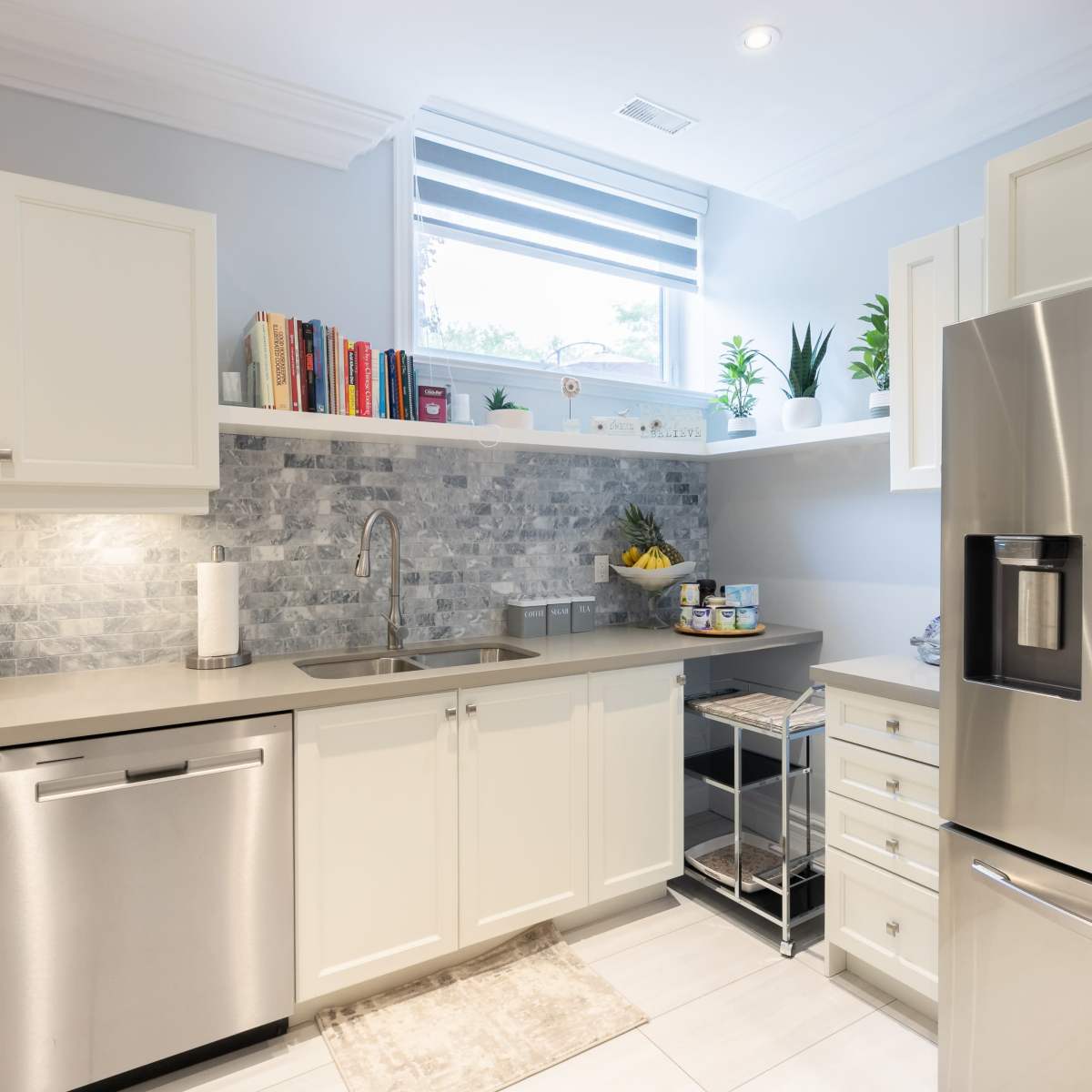 A modern kitchen features white cabinetry, a stainless steel refrigerator, and a window with sheer blinds, all arranged around a gray tile backsplash.