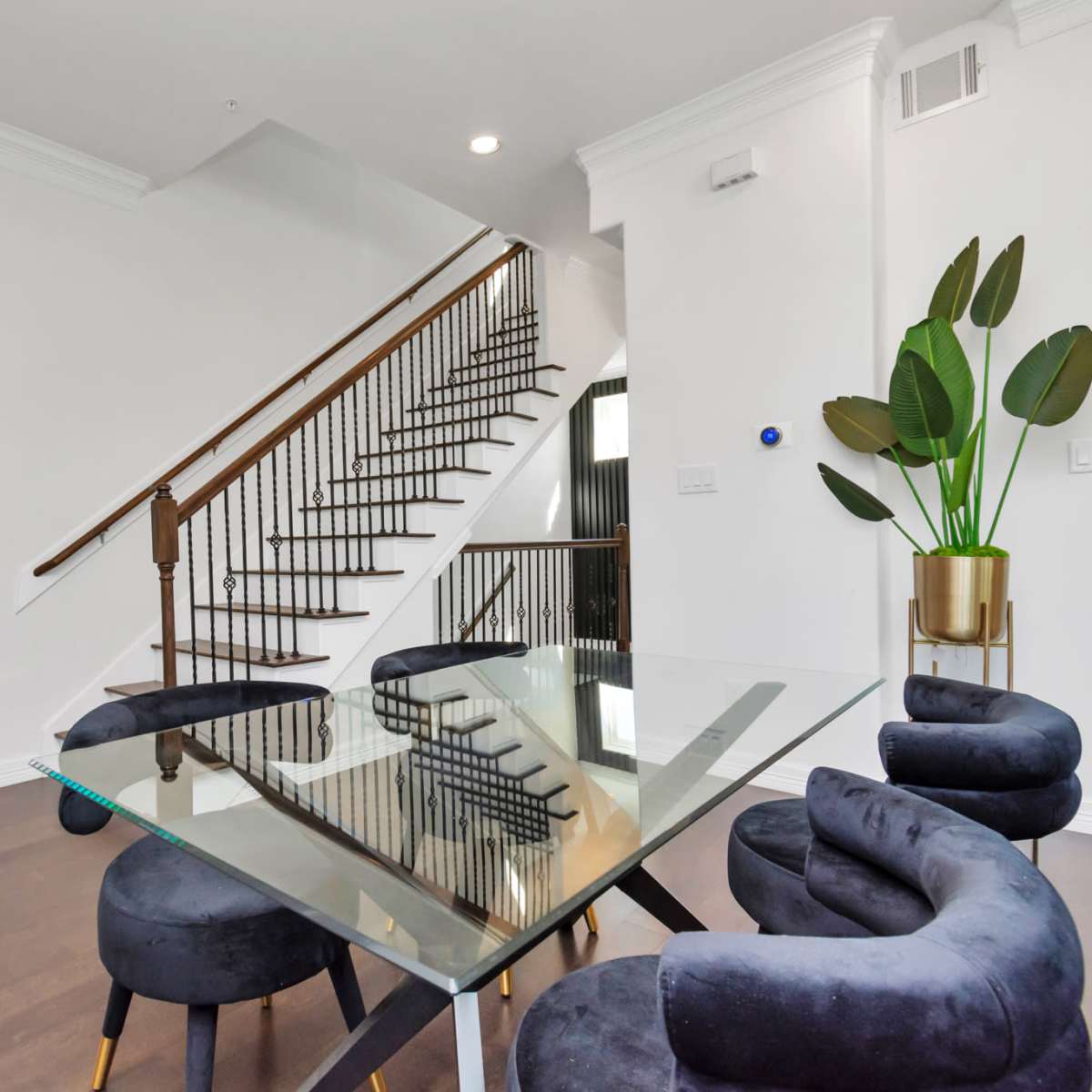 A modern dining area features a glass table surrounded by blue velvet chairs, with a staircase and a large mirror reflecting the space.