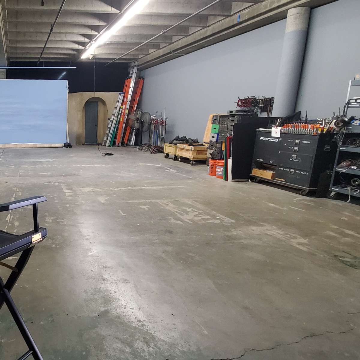 A vacant studio space featuring two director's chairs facing a backdrop, with equipment and props stored along the walls.