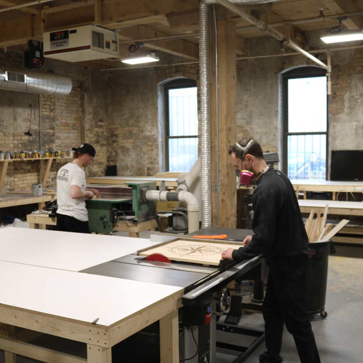 Two individuals are working in a woodshop, one operating a table saw and the other using a planer.
