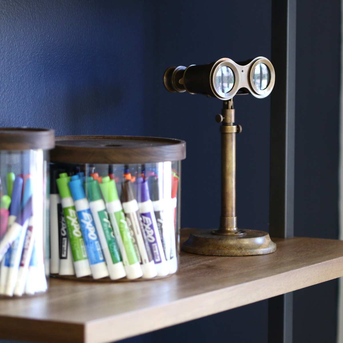 A wooden shelf displays two transparent containers filled with markers and a small vintage binoculars stand.
