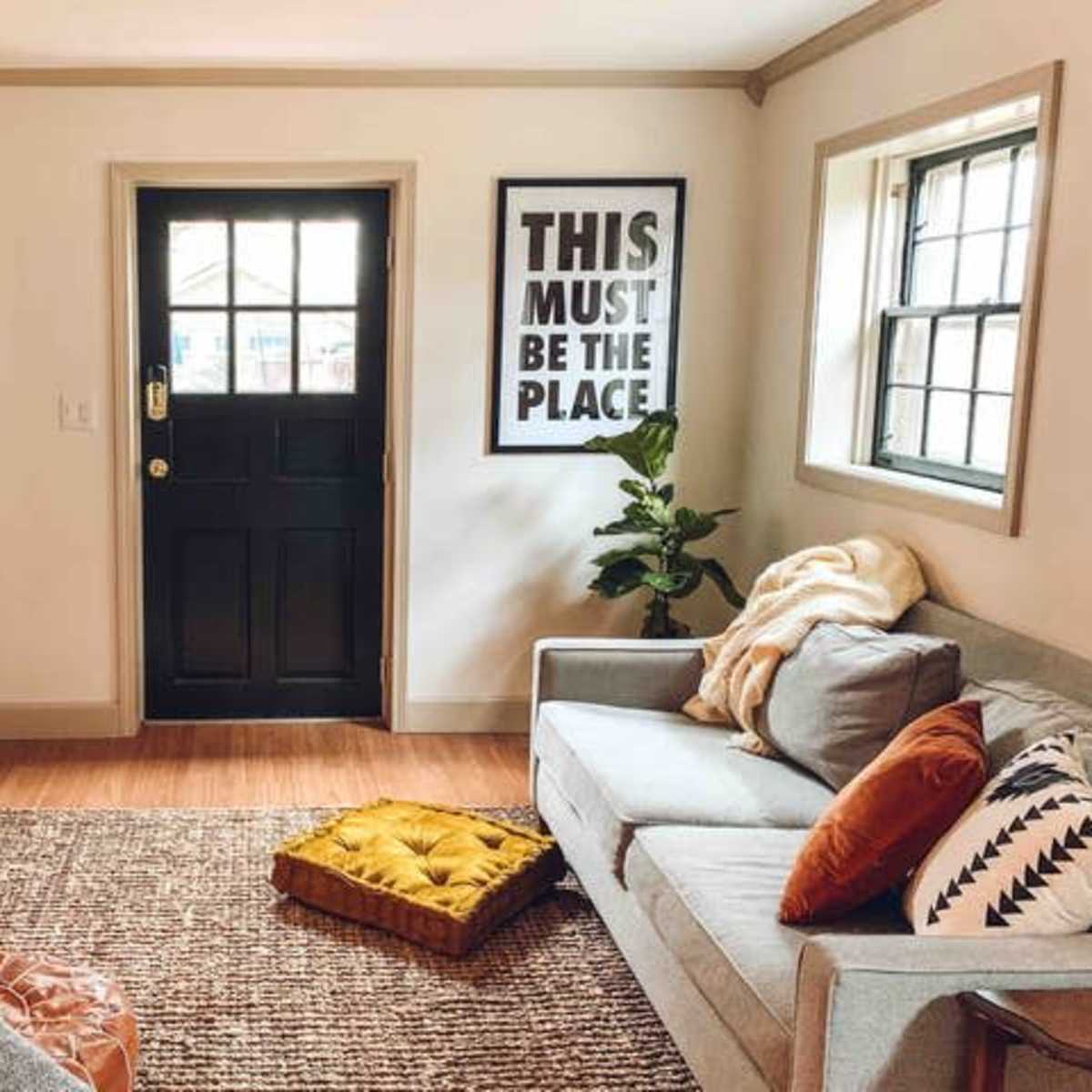 A living room features a gray sofa, an orange throw pillow, a yellow cushion on the floor, a flat-screen TV, and a door with a large framed poster beside it.