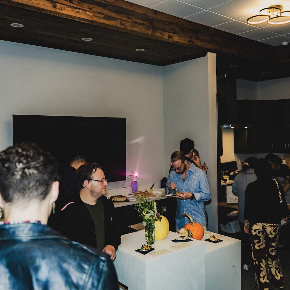 A group of people is gathered in a room with modern kitchen elements, engaging in conversation while examining displays on white pedestals.