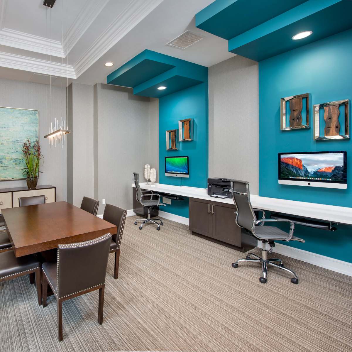A modern office space featuring a wooden dining table, dark chairs, a light fixture, and two workstations against teal walls, with artwork and decorative elements displayed.