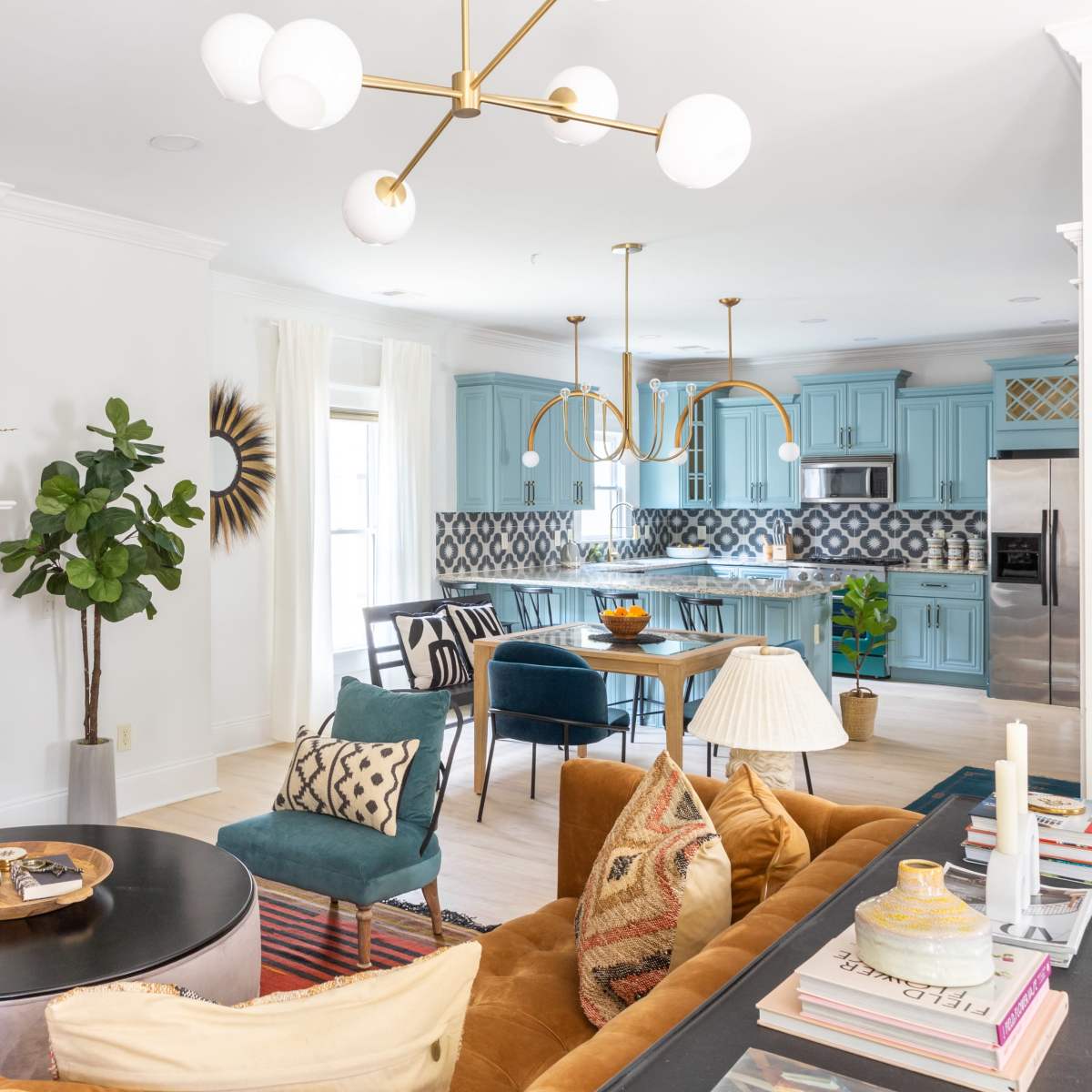 A modern living room with a large flat-screen TV above a fireplace, featuring a mix of colorful furniture, including a brown sofa and blue chairs, adjacent to a brightly colored kitchen area.