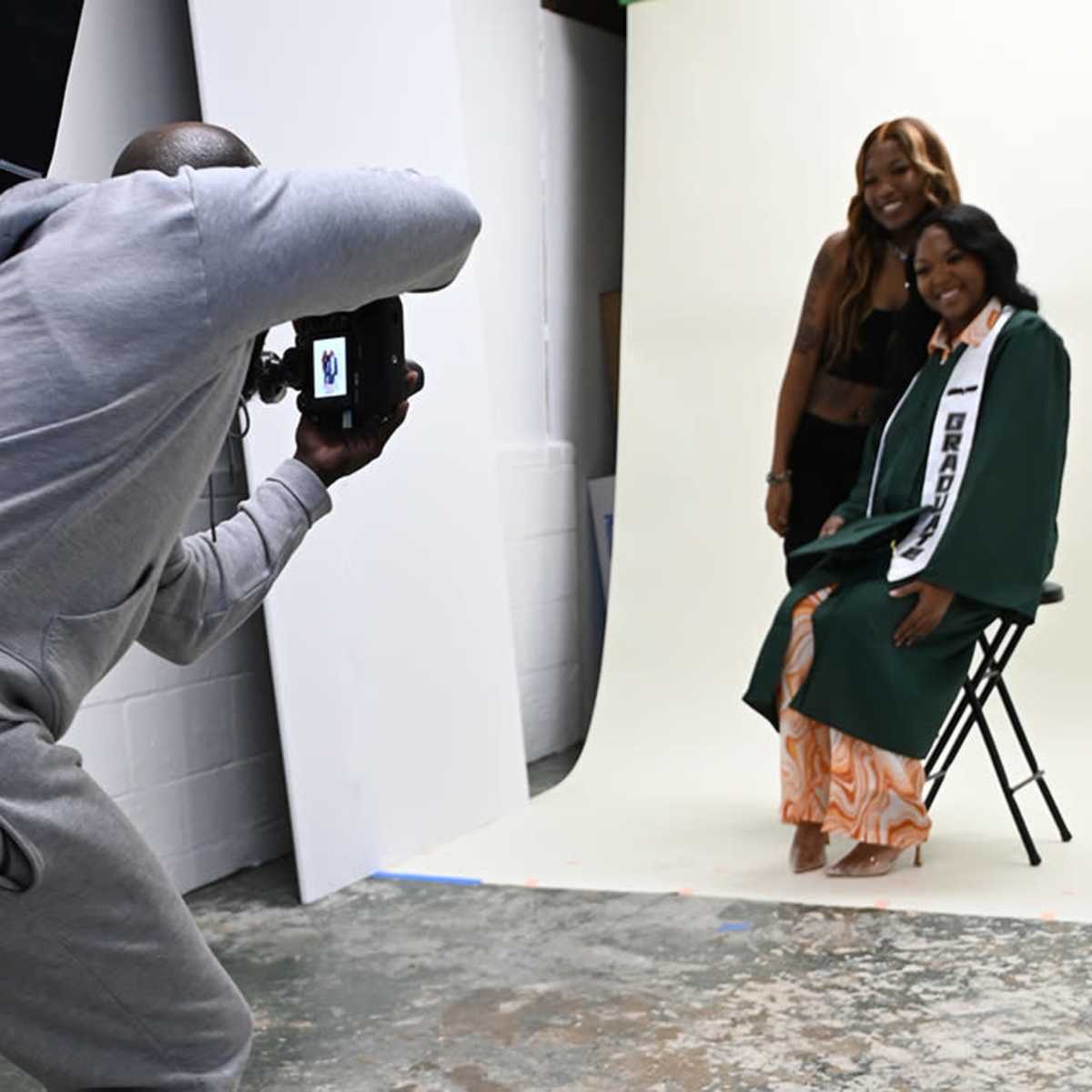 A photographer is taking a portrait of a woman in a graduation gown while another woman poses beside her in a studio.