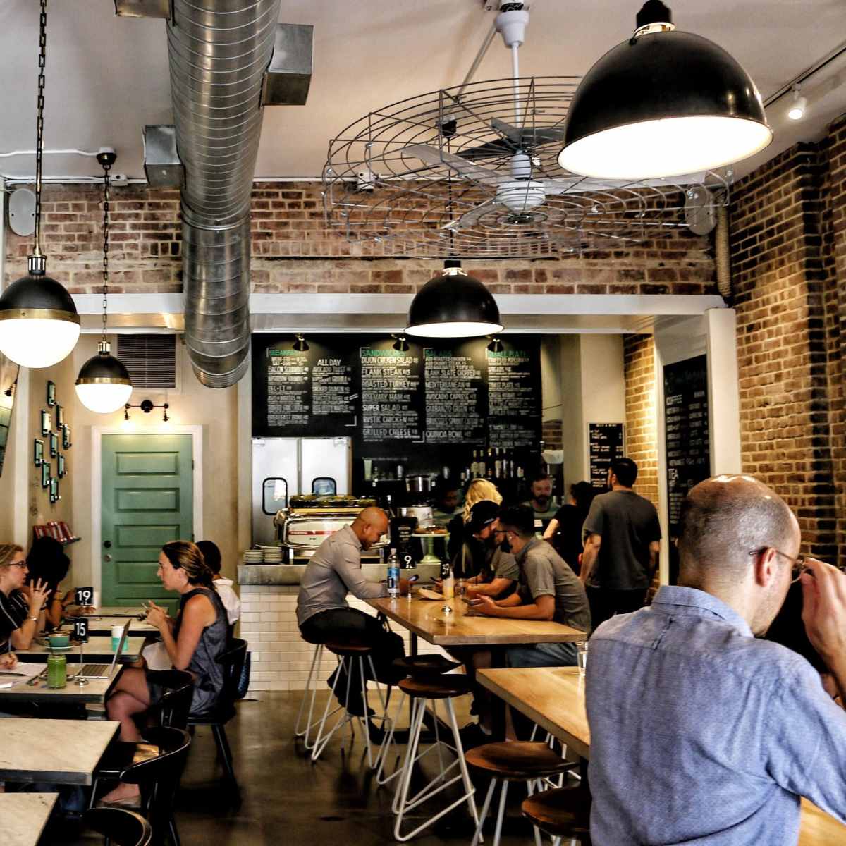 A busy café with a mix of patrons seated at various tables, exposed brick walls, and industrial-style lighting.