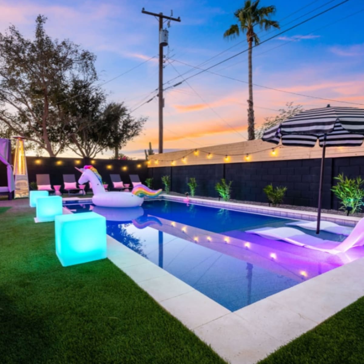 A backyard pool area with colorful lounge chairs, a floating inflatable unicorn, and a striped umbrella beside a modern swimming pool surrounded by lush greenery at sunset.