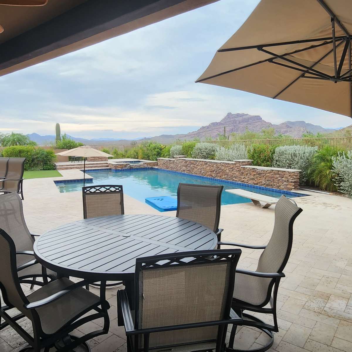 An outdoor patio with a round table and chairs, a swimming pool, and mountains in the background.