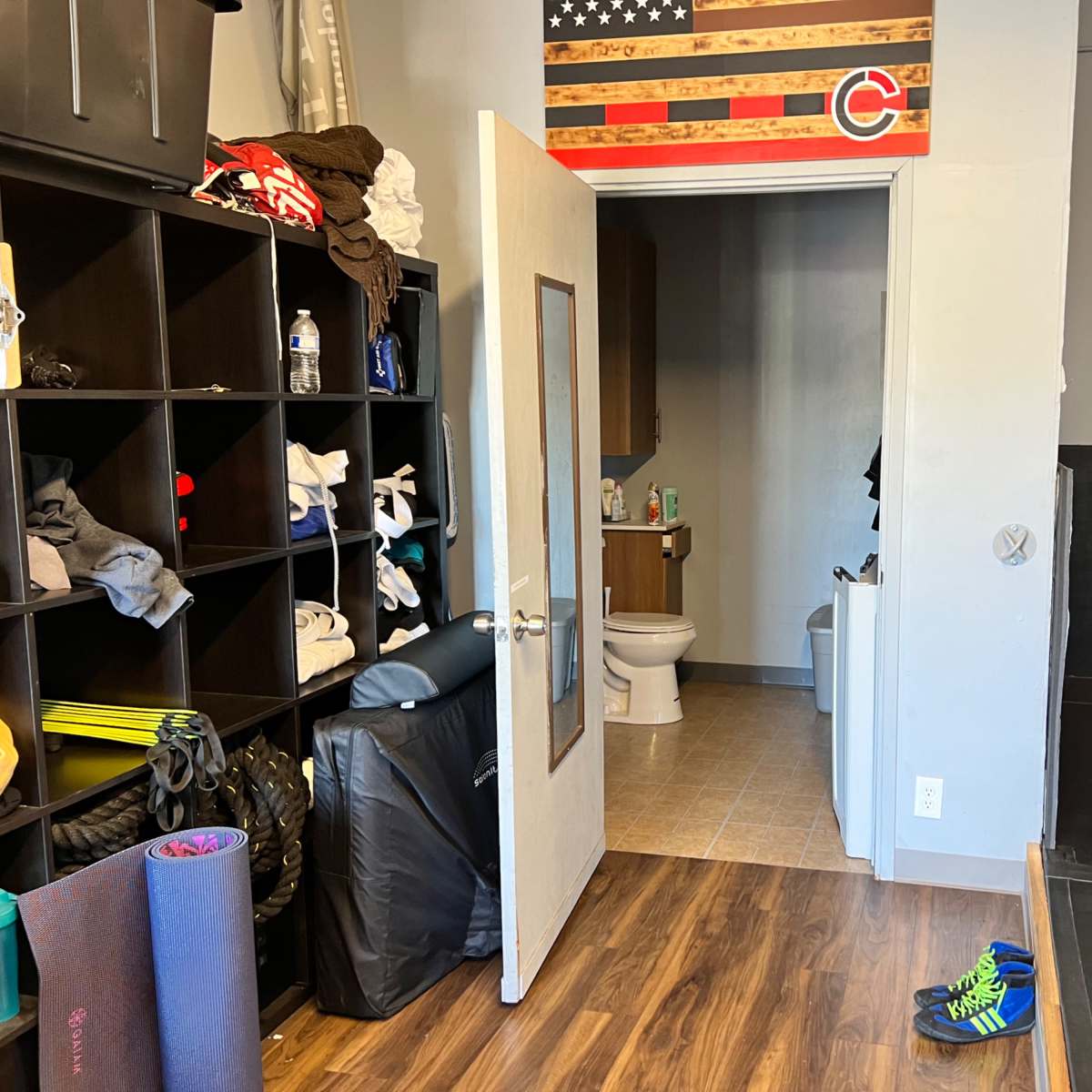 A room with a storage shelf filled with clothing, a partially open door leading to a restroom, and exercise equipment on the floor.