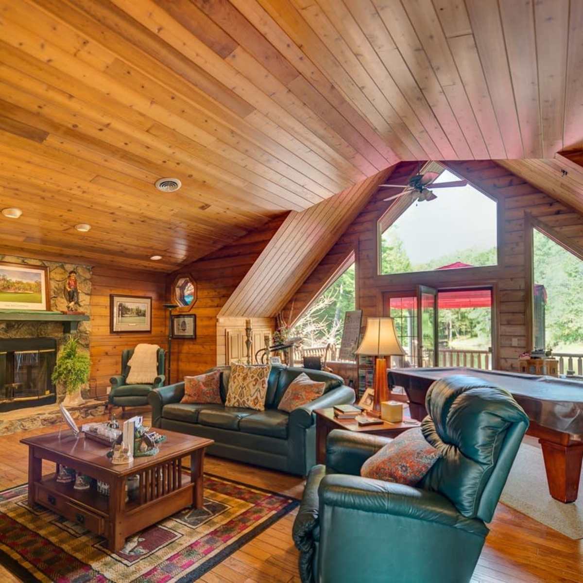 The living room features wooden walls, a stone fireplace, and large windows overlooking a deck.
