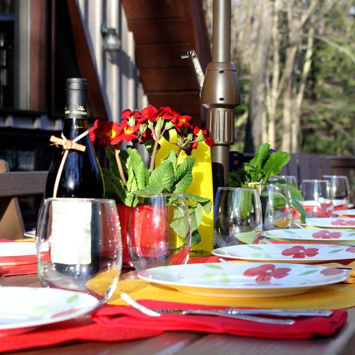 A long wooden table is set for a meal outdoors, featuring colorful plates, glasses, and a centerpiece of flowers, with a barbecue grill in the background.