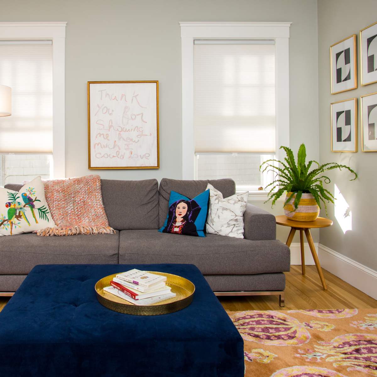 A cozy living room featuring a gray sofa with patterned throw pillows, a large blue ottoman, and framed artwork on the walls.