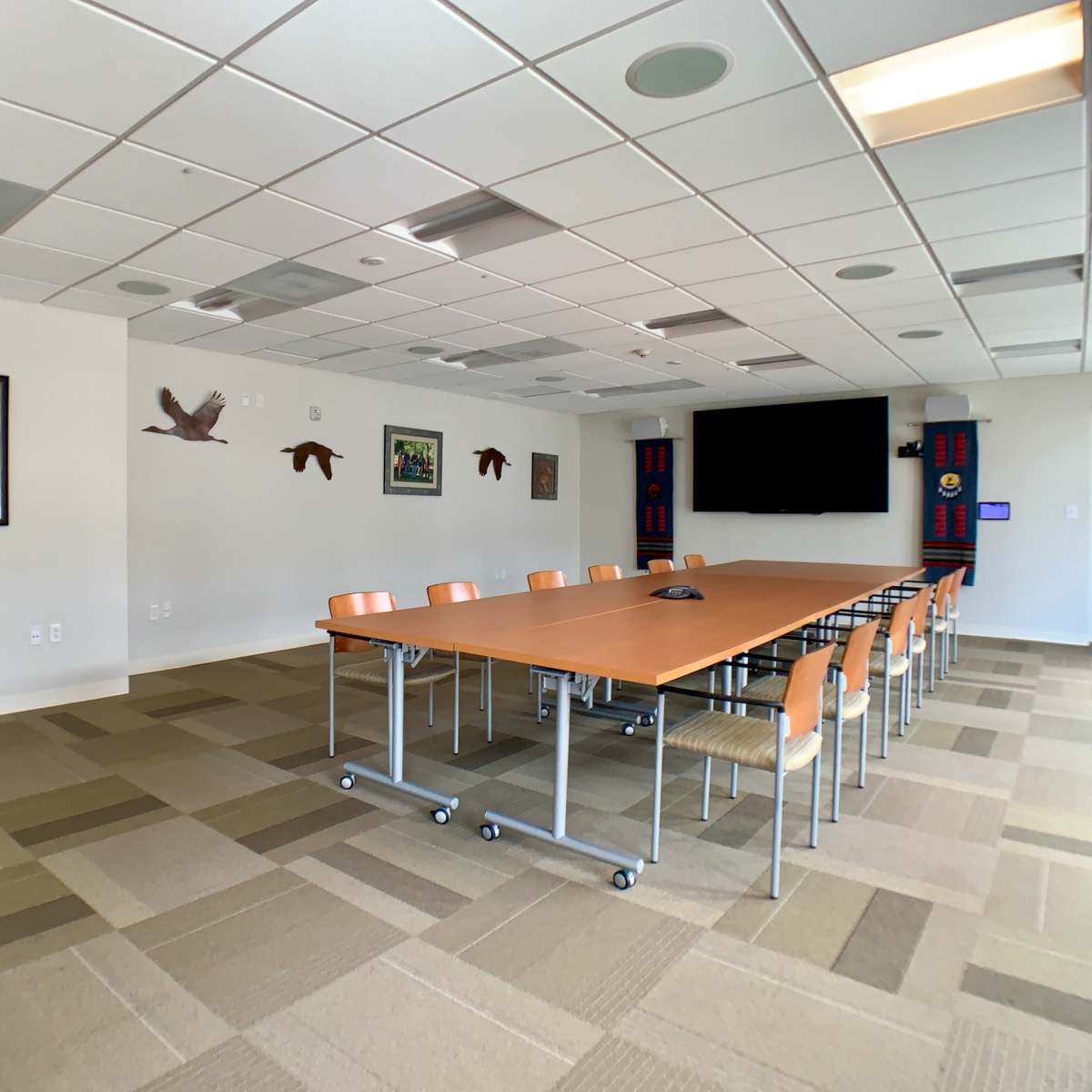 A large conference room features a long wooden table surrounded by chairs, with a wall-mounted screen and various framed artworks.