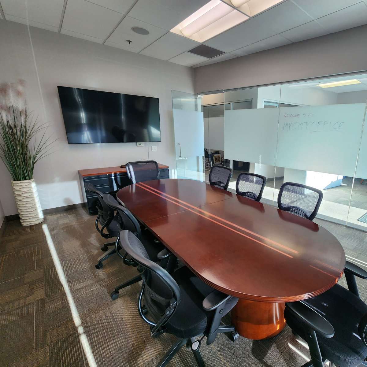 A conference room features a large wooden oval table surrounded by black mesh ergonomic chairs, with a whiteboard and a TV on the wall.
