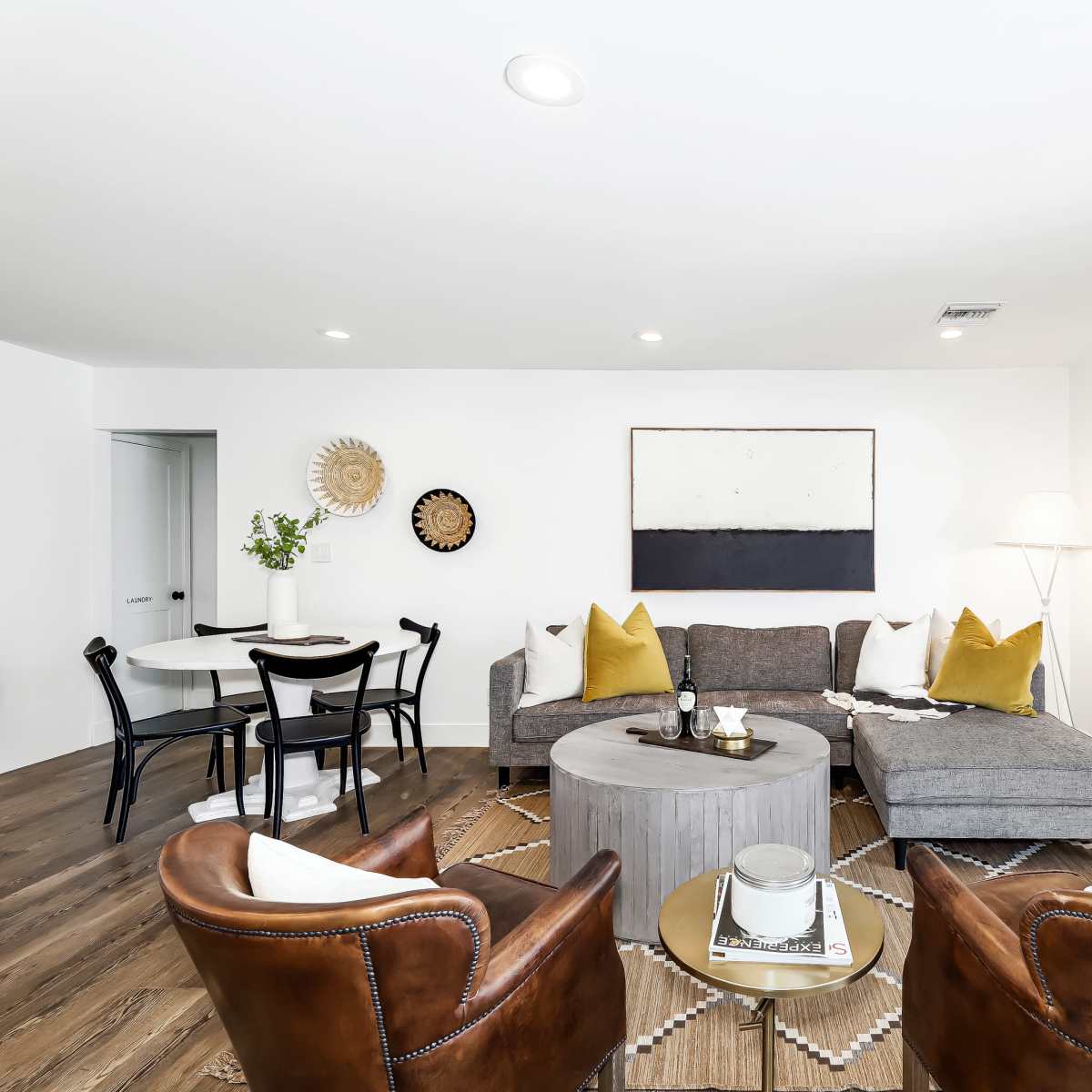 A modern living room featuring a gray sectional sofa, brown leather armchairs, a round coffee table, and a wall art piece, with a dining area visible in the background.
