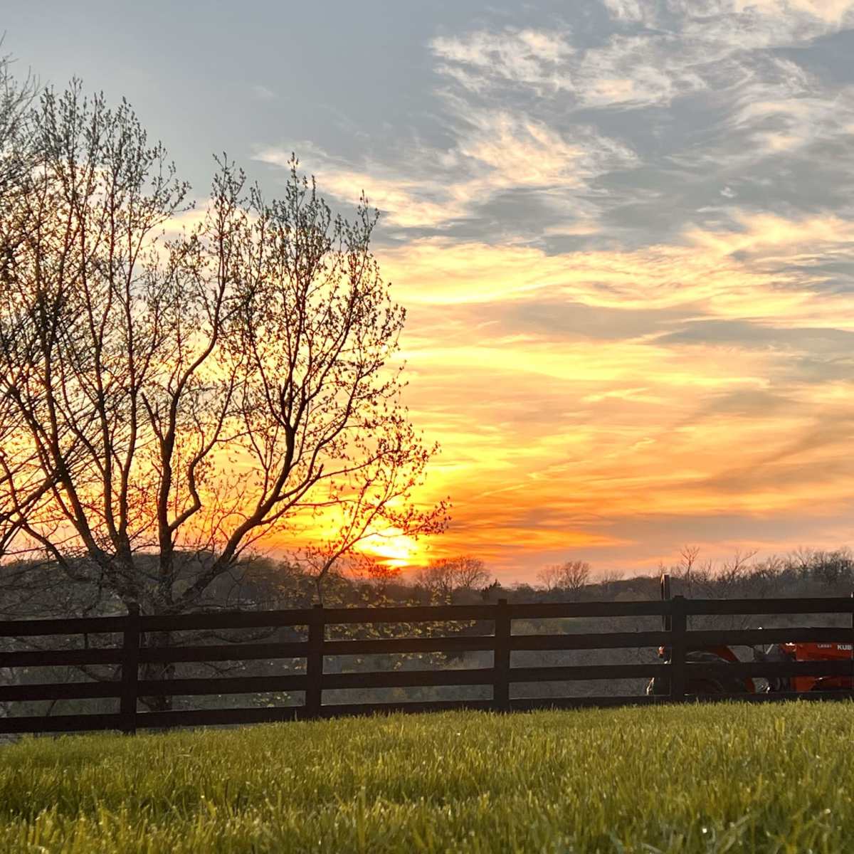 The sunset casts a warm glow over a fenced landscape with trees and a tractor in the foreground.