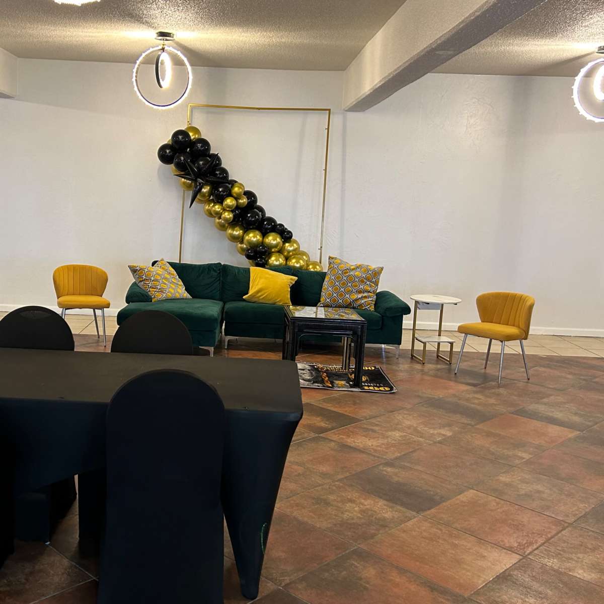A modern event space featuring a green sofa with decorative pillows, a black and gold balloon arrangement, a table covered with a black cloth, and yellow chairs.