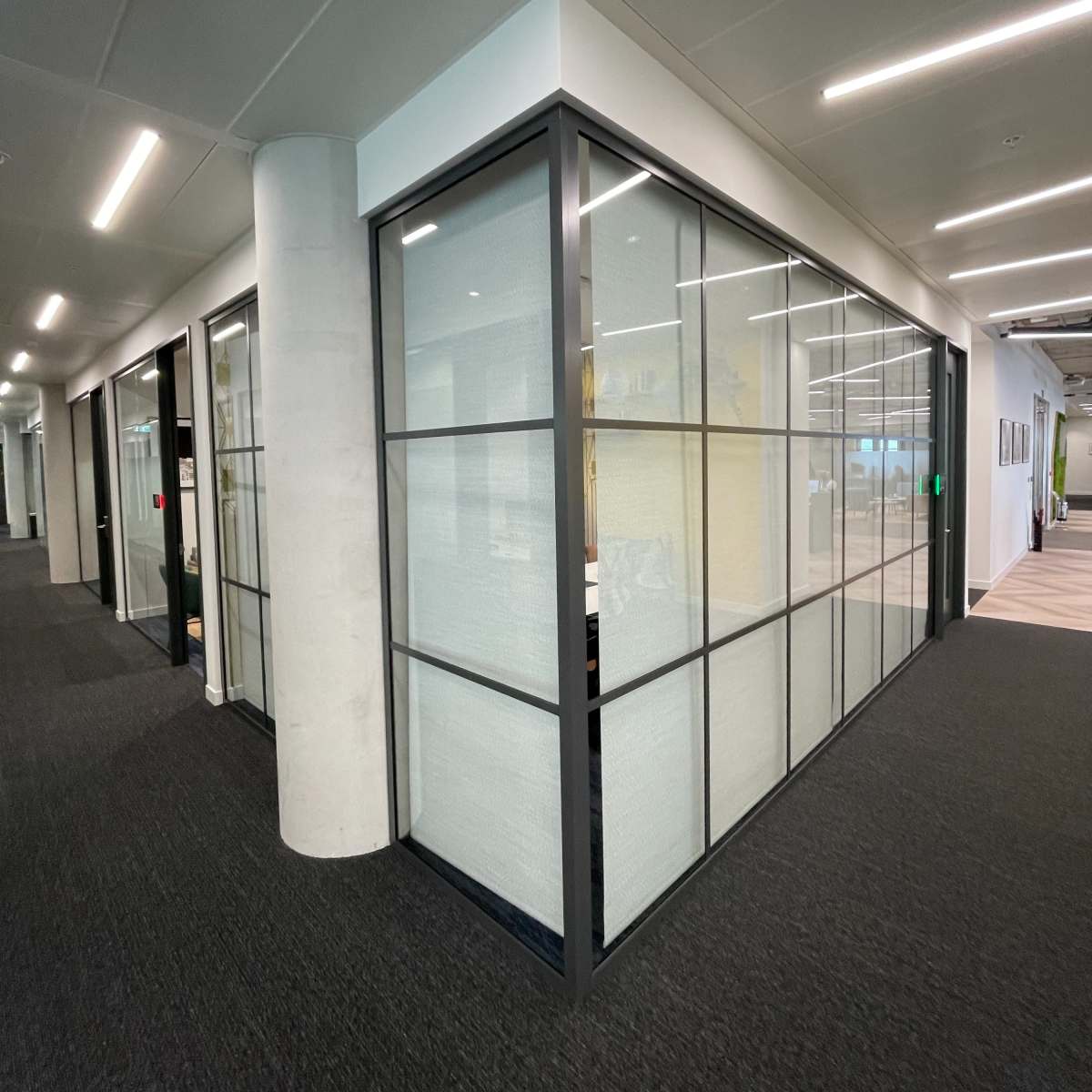 A modern office space featuring glass-walled meeting rooms and a dark carpeted flooring.
