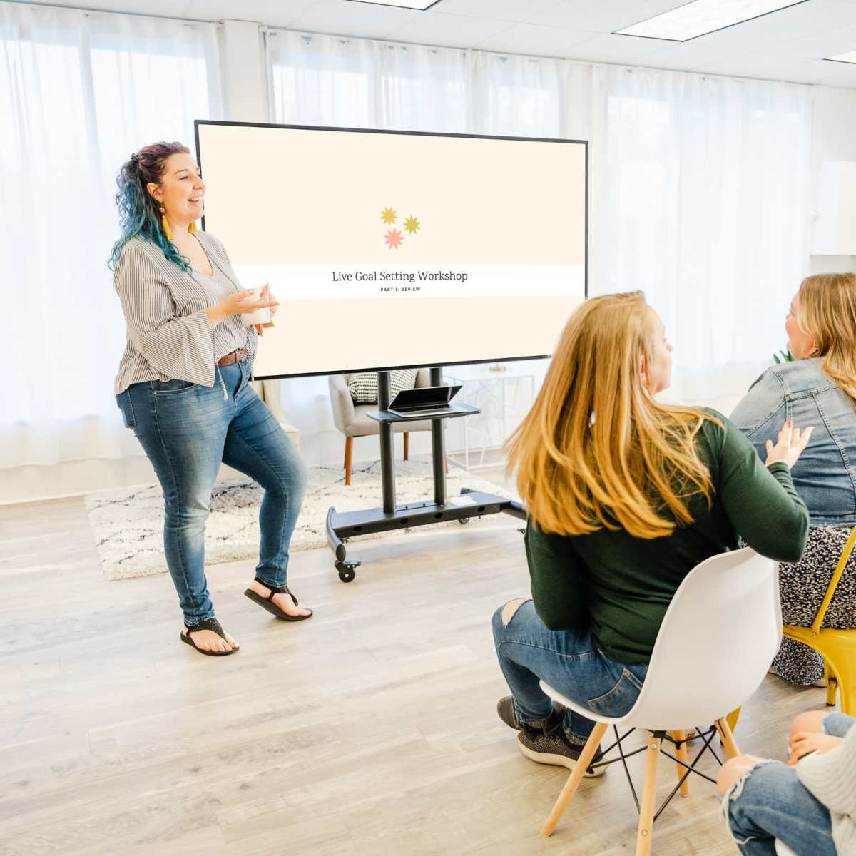 A woman with blue hair leads a goal-setting workshop in front of an audience seated on colorful chairs.