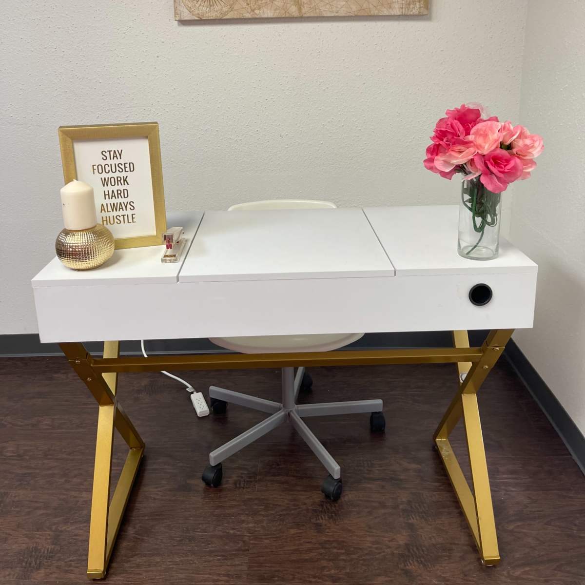 A white desk with a gold frame, accompanied by a chair, a framed motivational quote, a flower vase, and a candle, is set against a wall with a world map.