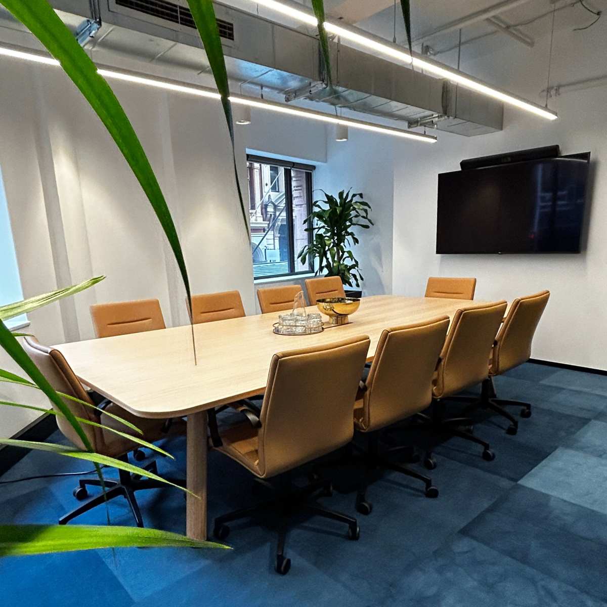 A spacious conference room features a large wooden table surrounded by ergonomic chairs, with plants and a wall-mounted screen in the background.