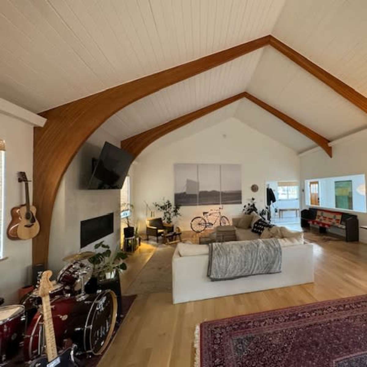 A spacious living room with a high, vaulted ceiling featuring exposed wooden beams, a large sectional sofa, a dining area, and various decorative elements including musical instruments and plants.