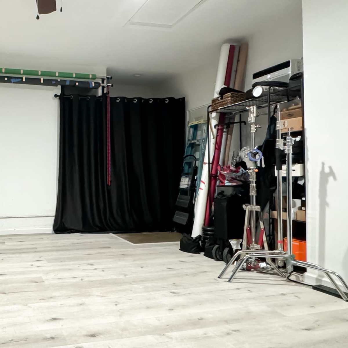 A minimalist indoor space with light wooden flooring, black curtains on one side, and various equipment and storage items against the wall.