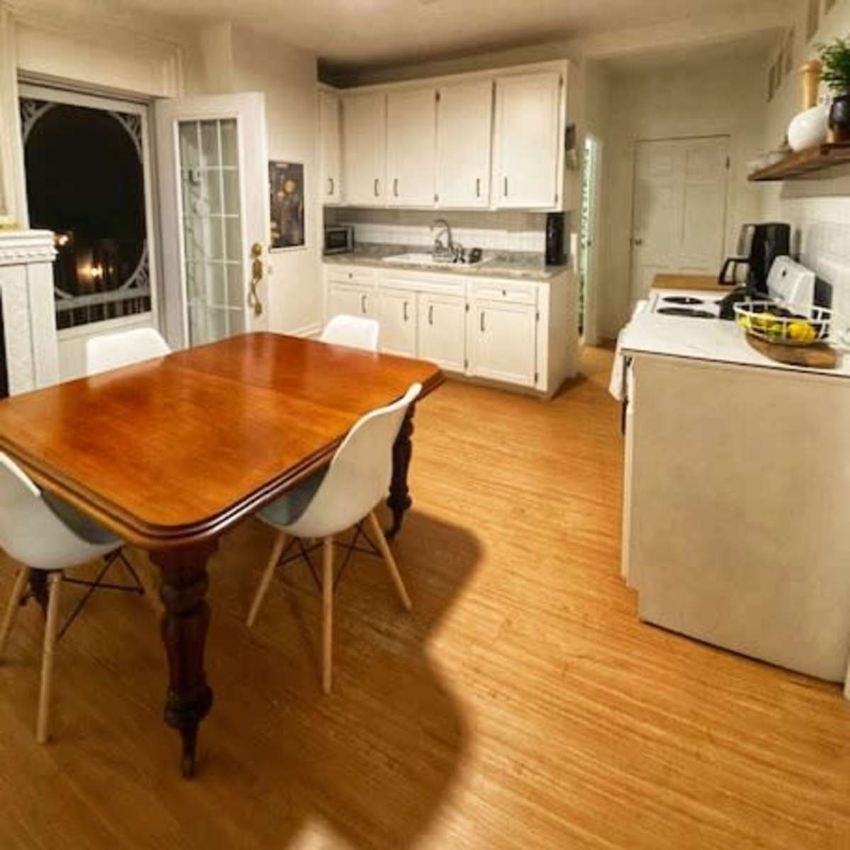A kitchen with a wooden dining table and white chairs, featuring a stove, refrigerator, and cabinets in a well-lit space.