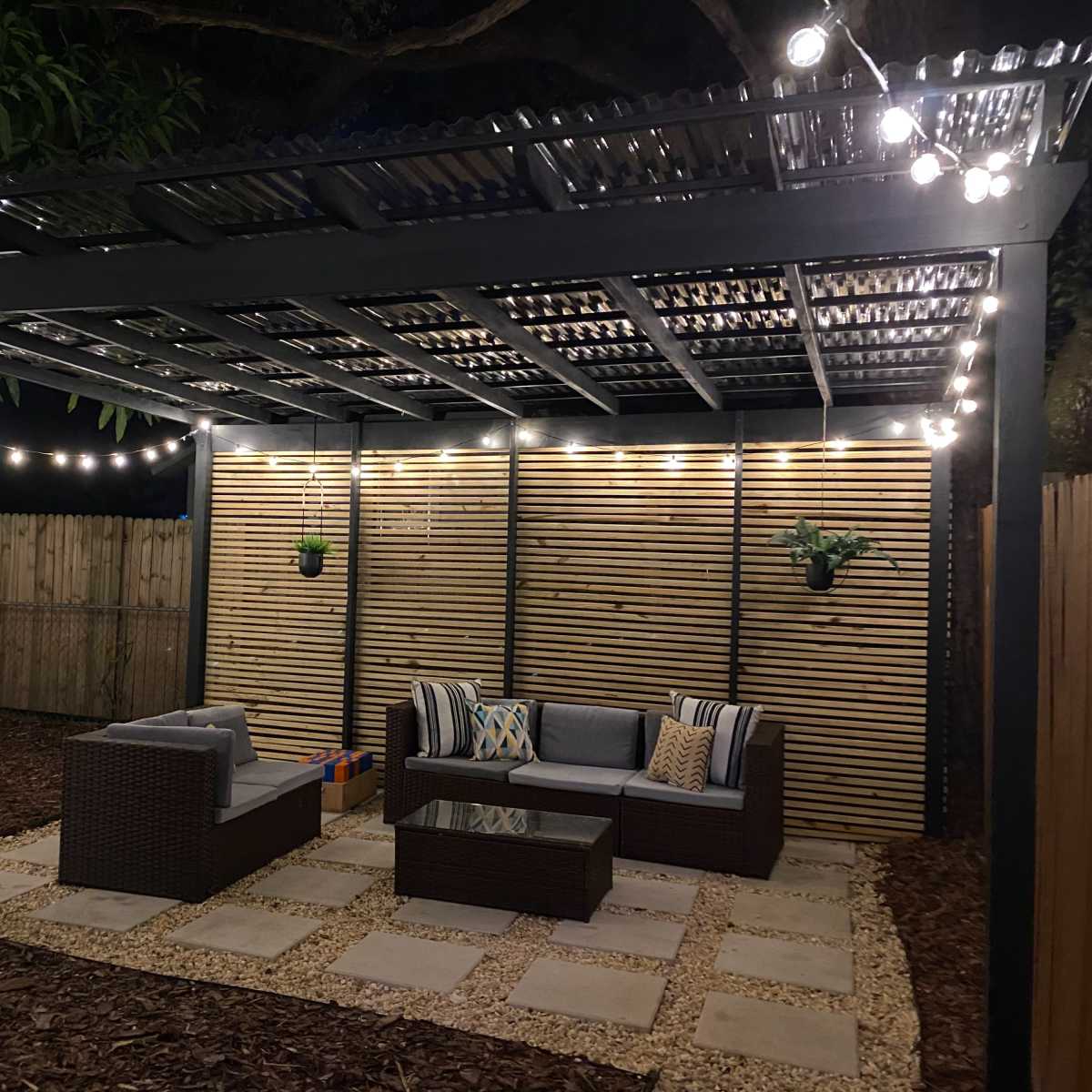 A patio area features a modern pergola with string lights, surrounded by landscaping and furnished with a seating arrangement and coffee table.