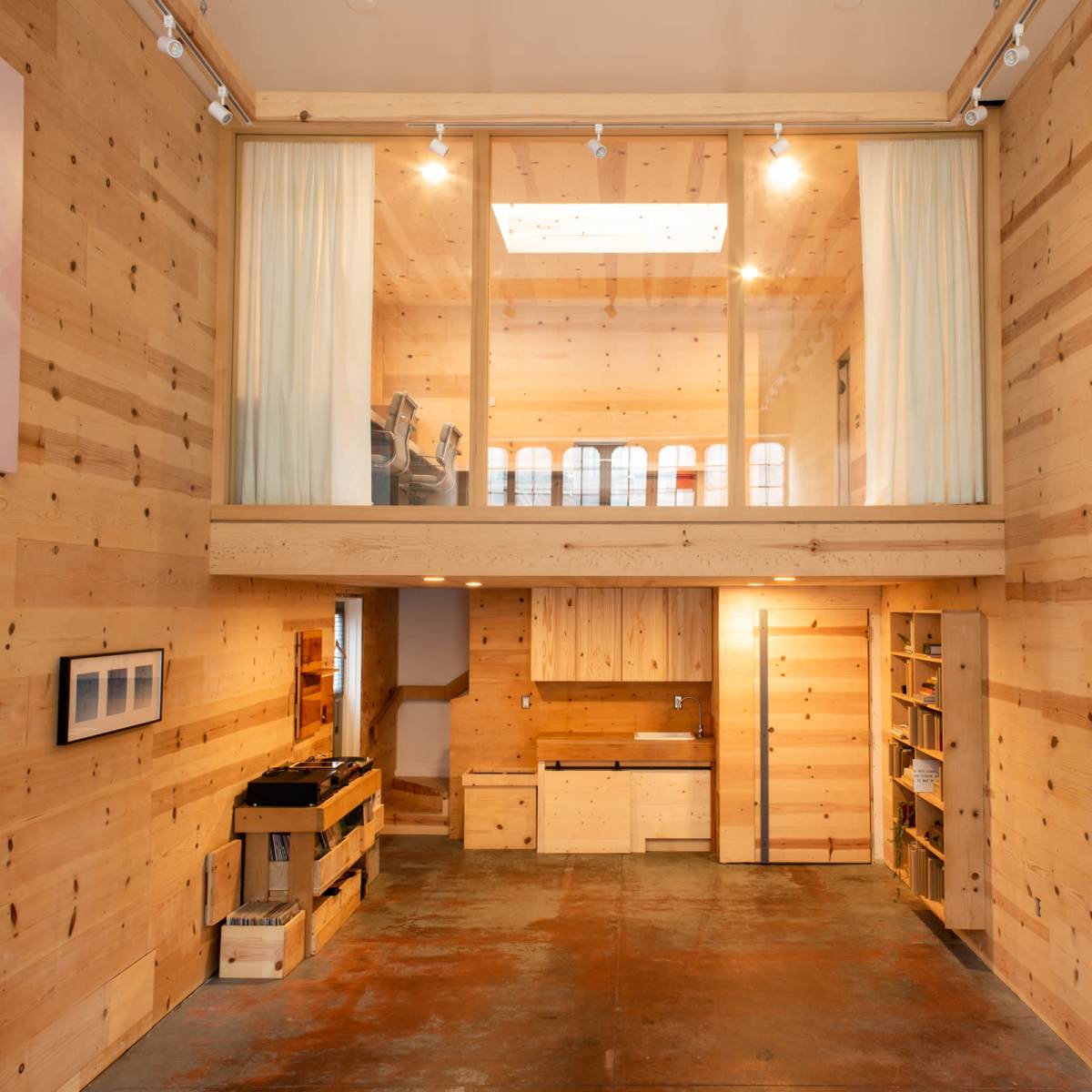 A spacious interior of a wooden room with a mezzanine level, featuring minimalist furniture, large windows, and polished concrete flooring.