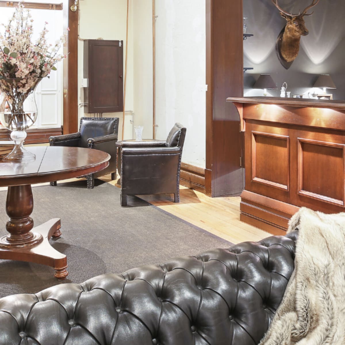 A reception area featuring a wooden front desk, a round dining table, leather chairs, and a decorative taxidermy deer head on the wall.