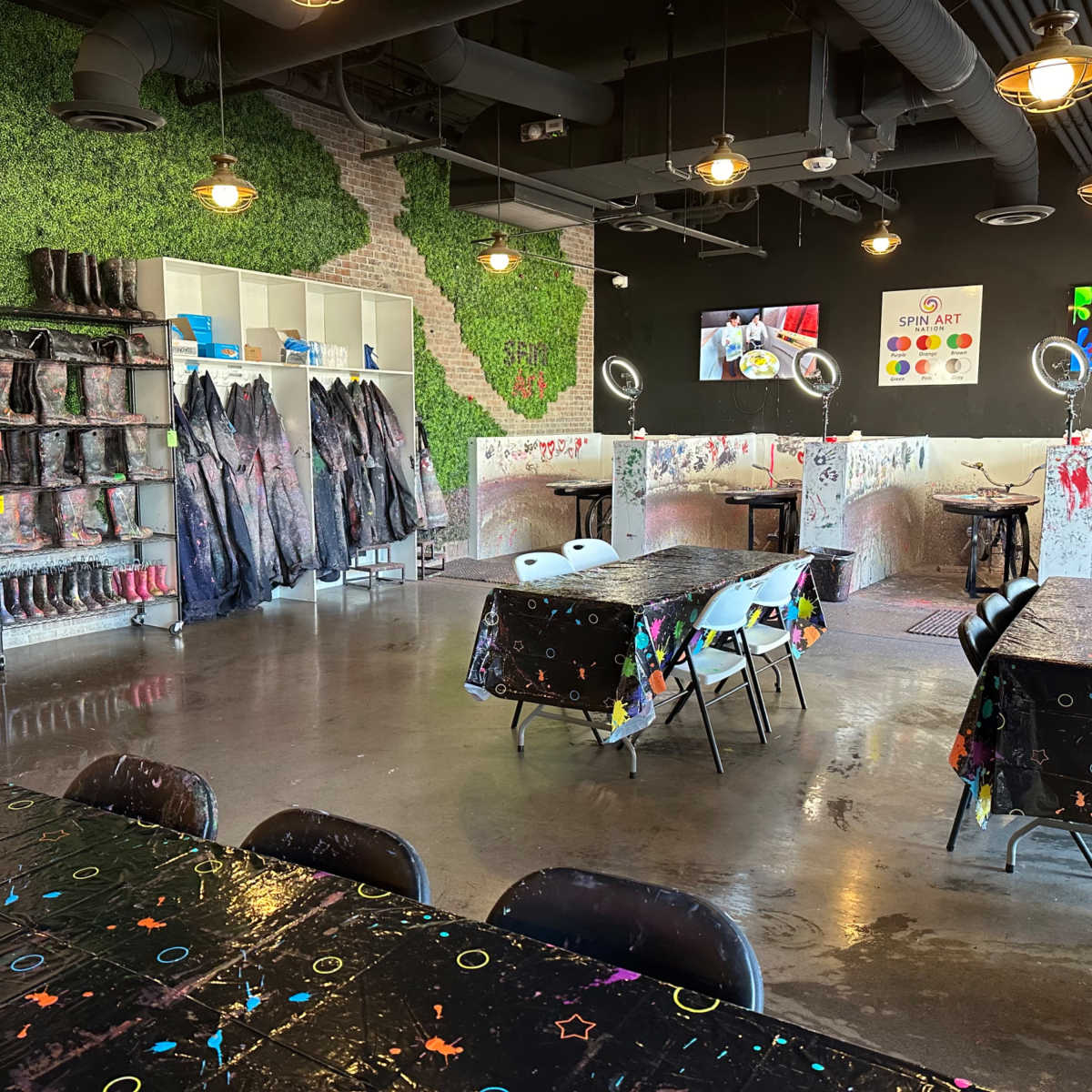 An art studio with tables covered in colorful tablecloths, black chairs, and a wall decorated with green foliage, alongside shelves of paint supplies and protective gear.