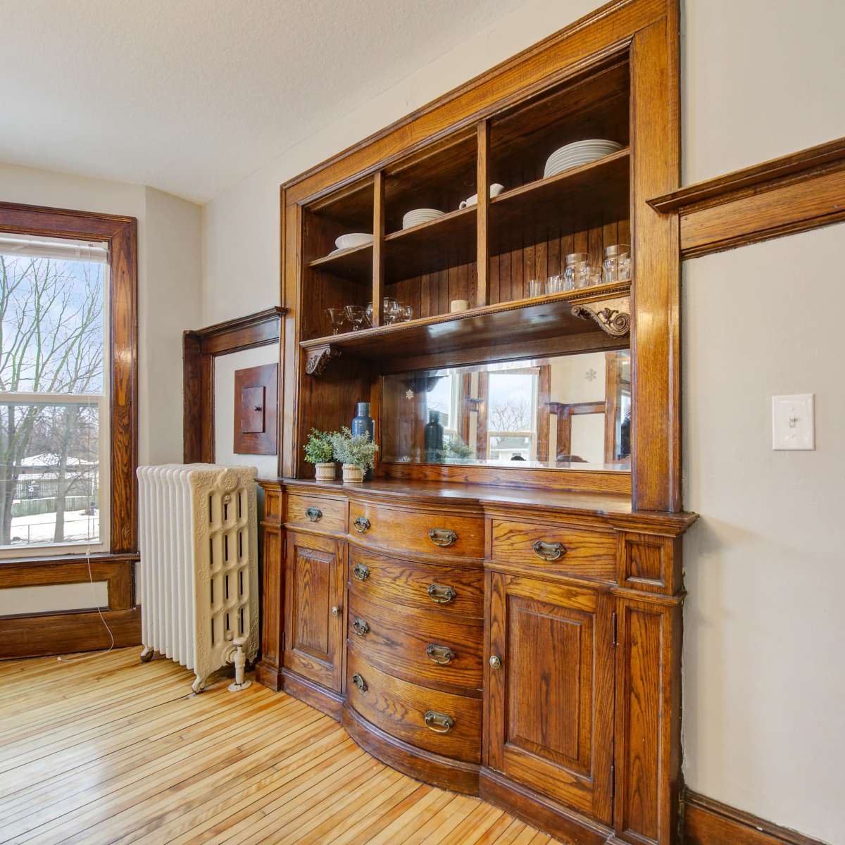 A wooden sideboard with glass shelves, situated in a room featuring wooden trim and large windows.