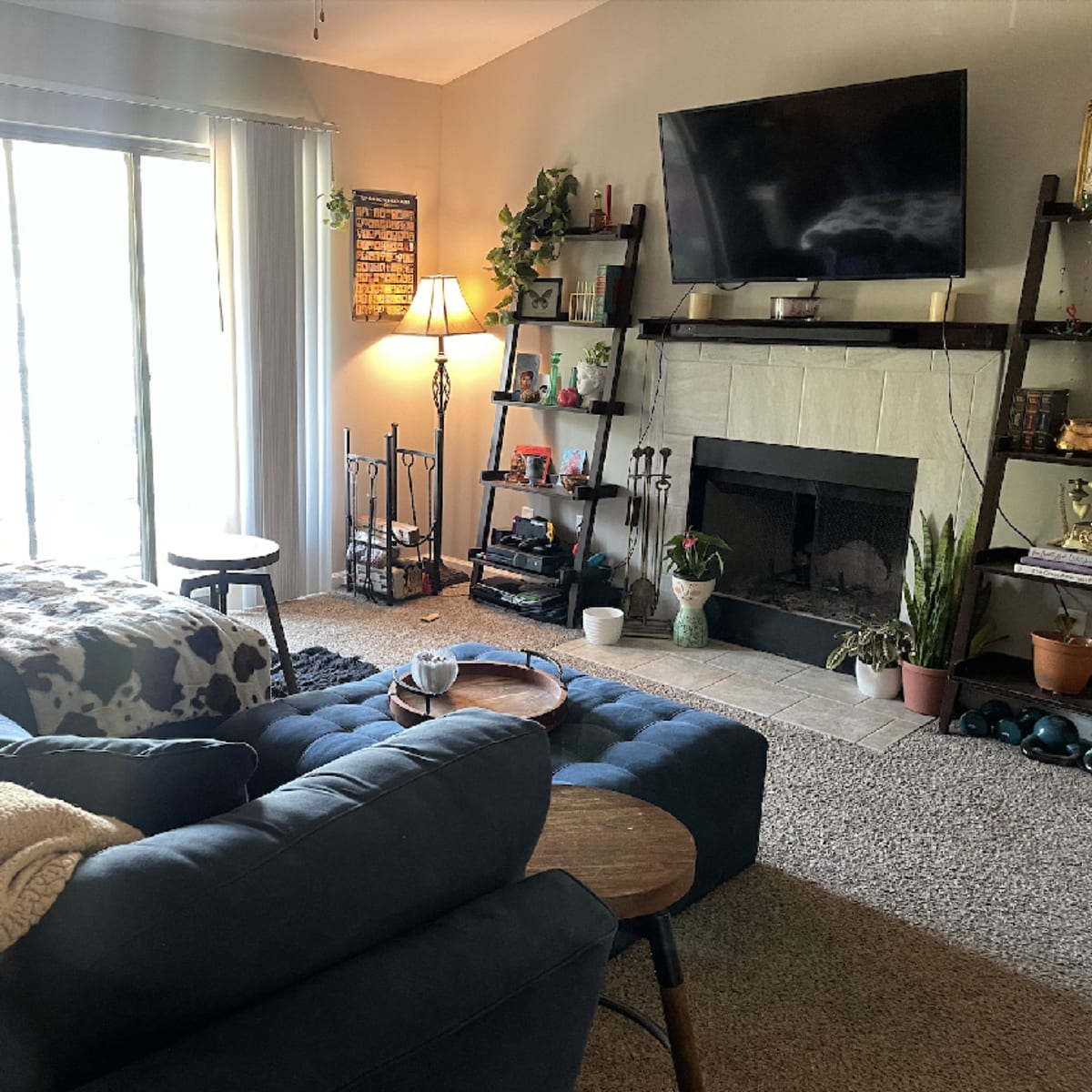 A cozy living room with a blue sectional sofa, a coffee table, and a fireplace surrounded by shelves and plants, illuminated by natural light from a sliding glass door.