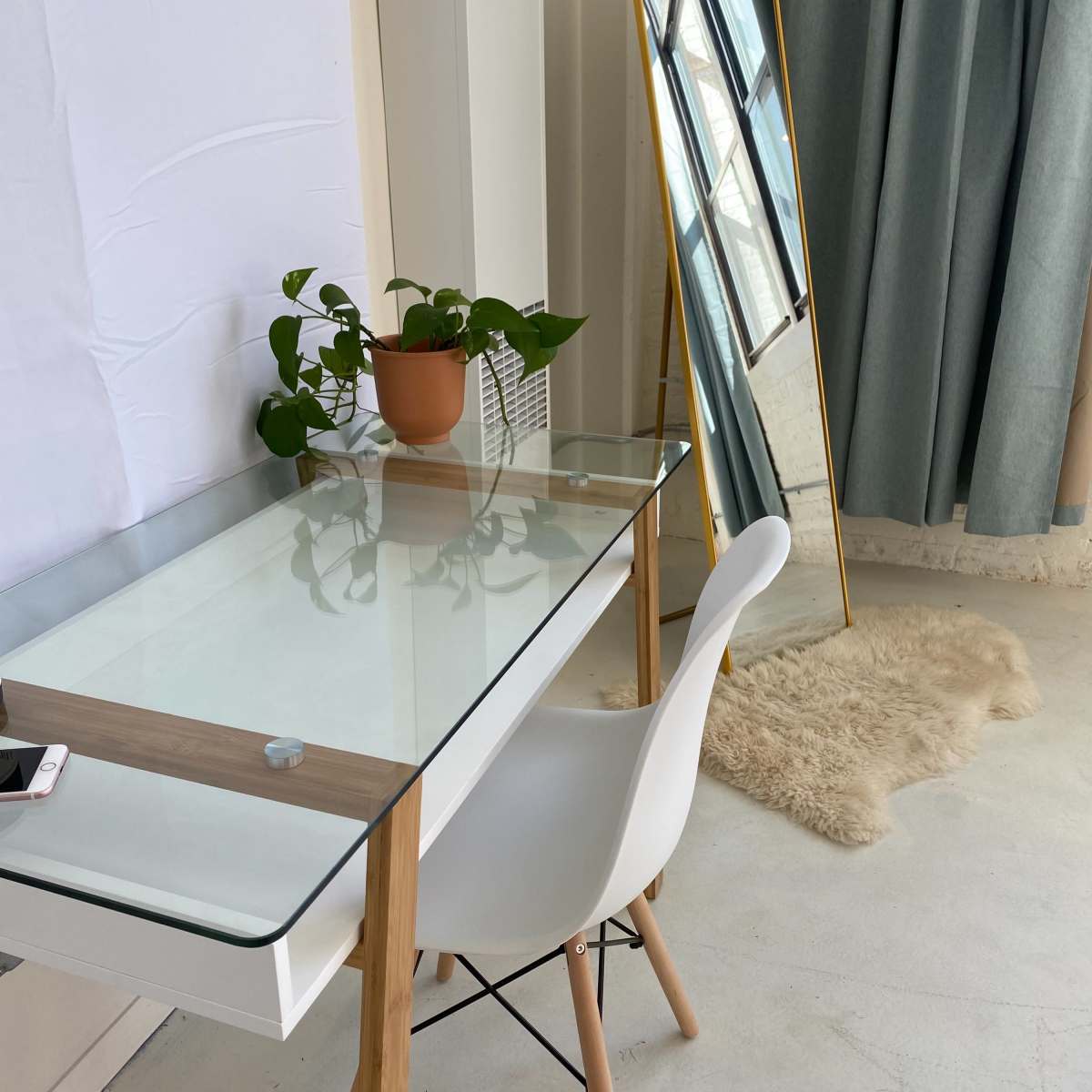 A clear glass desk with a wooden frame is positioned beside a tall gold-framed mirror and a potted plant on a white wall backdrop.