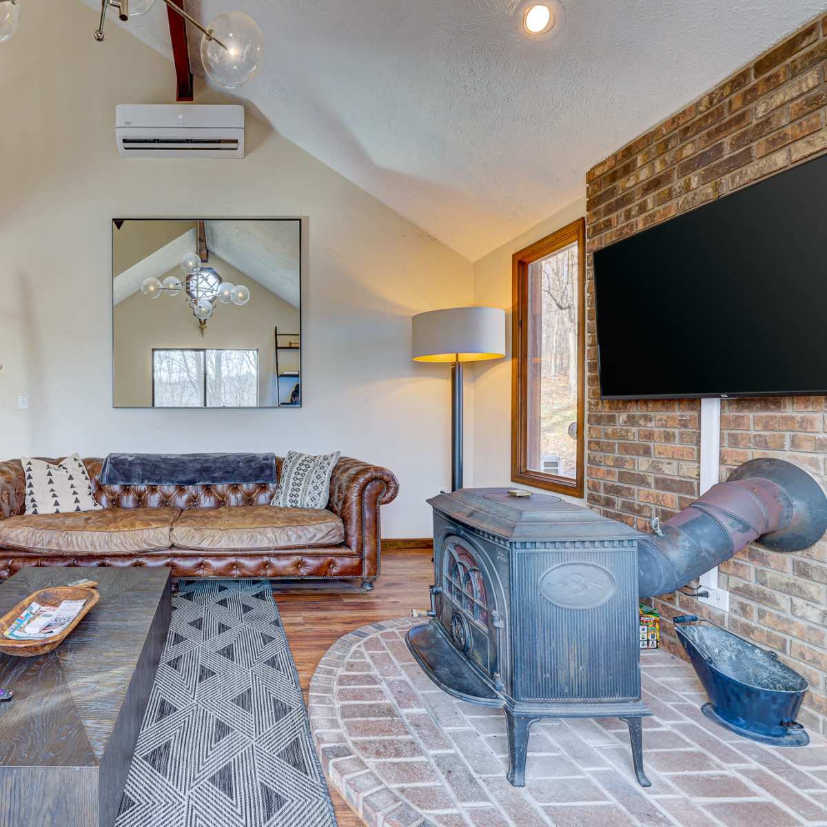 A cozy living room featuring a leather sofa, a wooden coffee table, a brick wall with a mounted TV, and a cast iron stove.