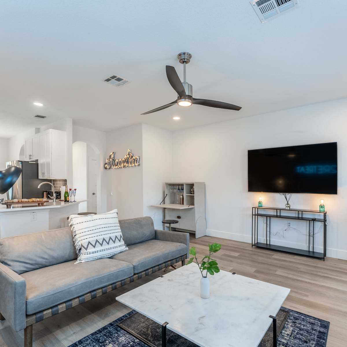 A modern living room with a gray sofa, a coffee table, and a wall-mounted television, arranged in a bright and open space that connects to a dining area.