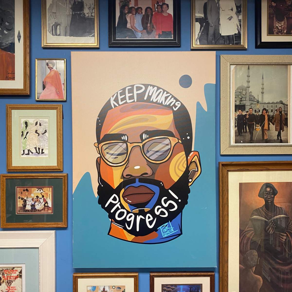 A colorful mural of a man with sunglasses and the phrase "KEEP making Progress!" surrounded by a variety of framed photographs on a blue wall.