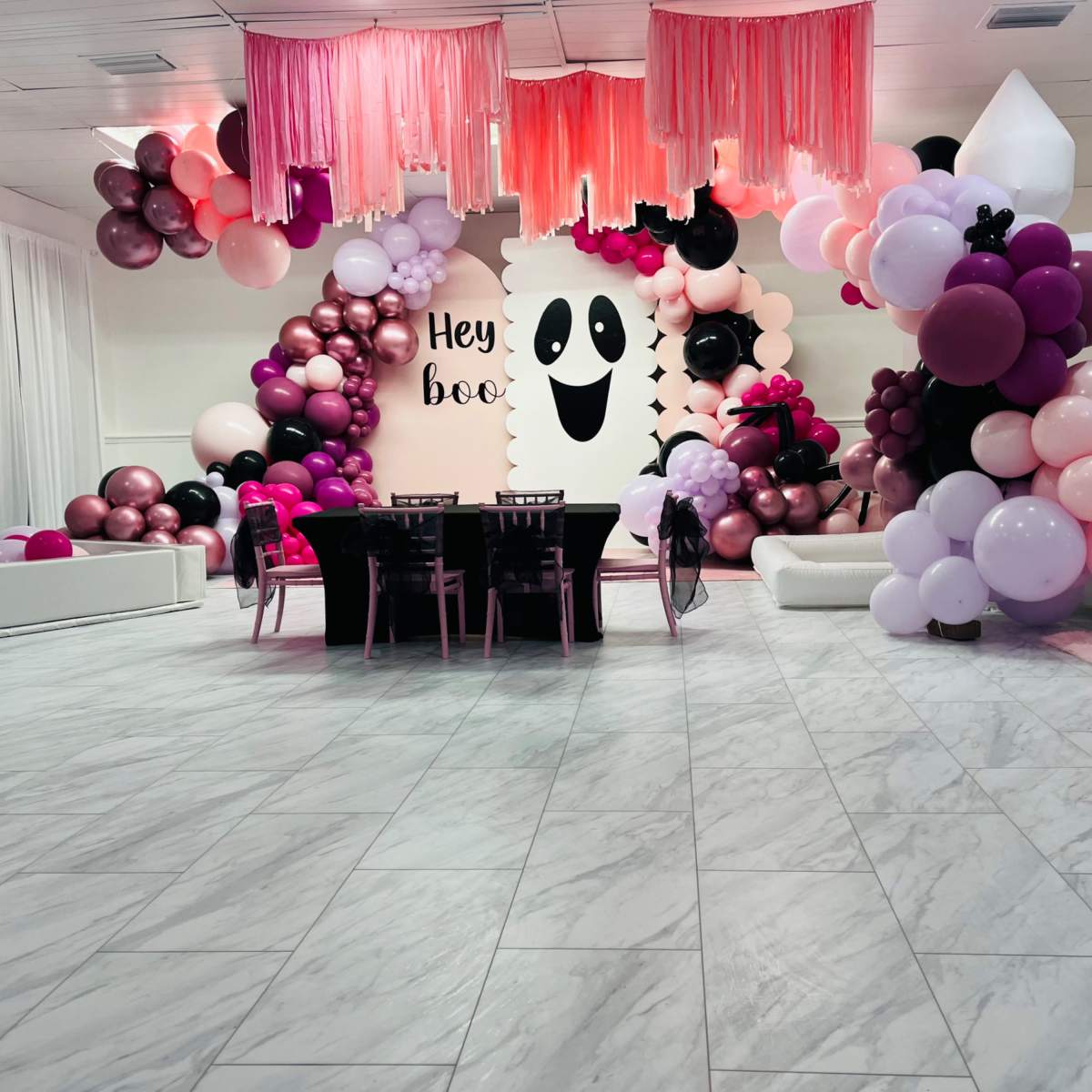 A decorated event space featuring a table set for guests, surrounded by an assortment of balloons in various colors and a wall with a ghost-themed design that reads "Hey boo."