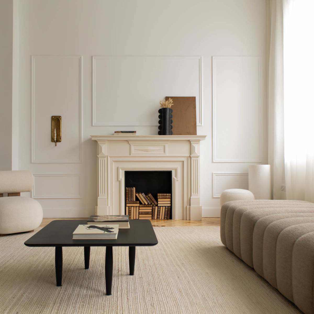 A modern living room features a light-colored sofa, a black coffee table, and a minimalist fireplace, with a large window allowing ample natural light.