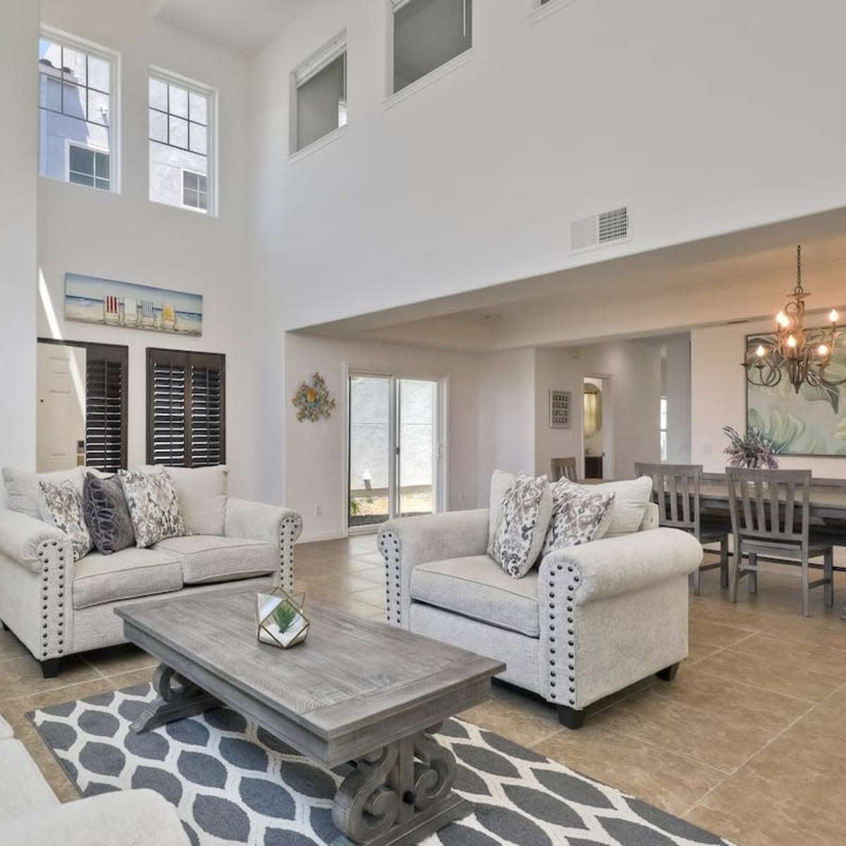 A bright, airy living room features two beige armchairs, a gray coffee table, and a beige sofa, with large windows illuminating the space.