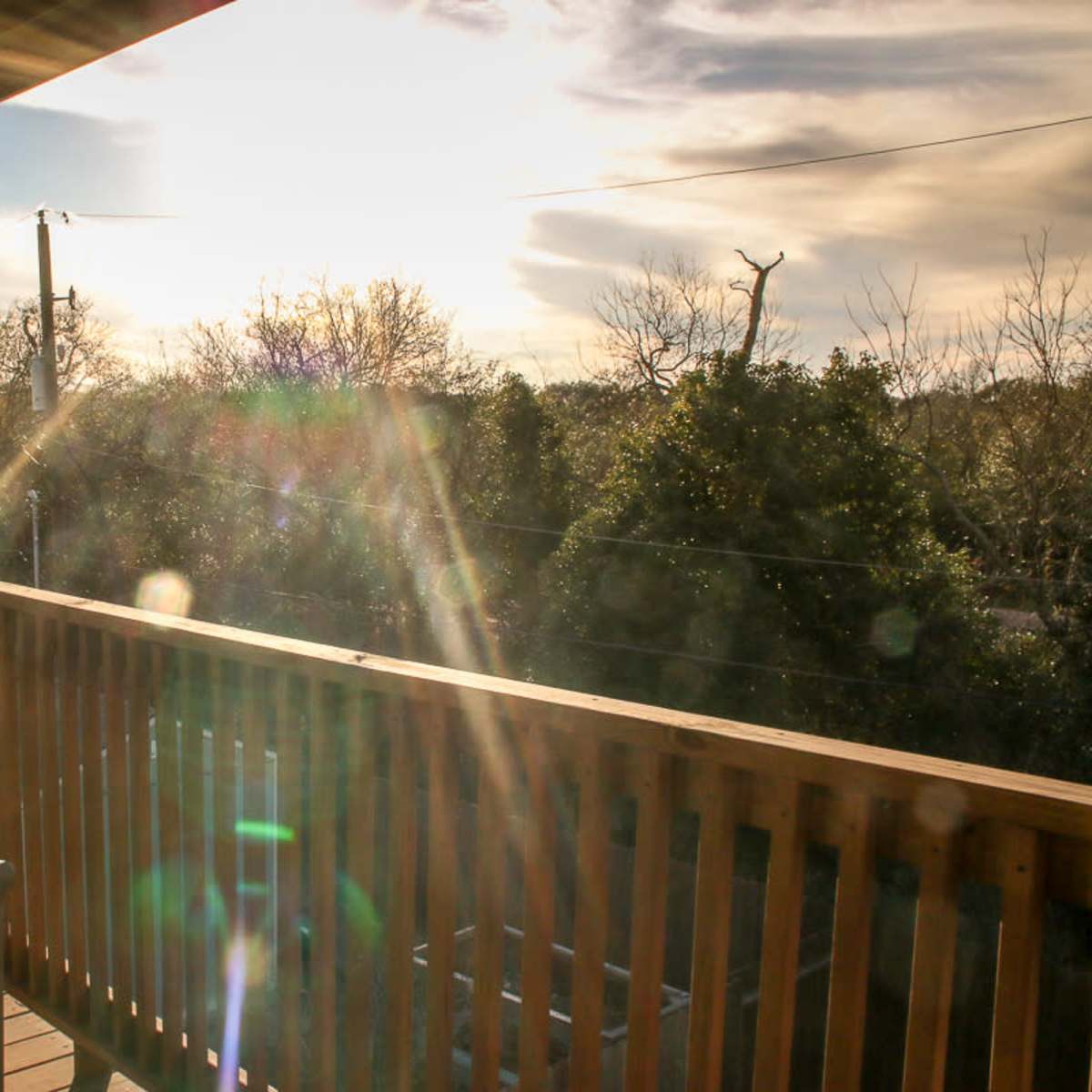 A wooden balcony with a bench, overlooking a landscape with trees and power lines under a bright sky.