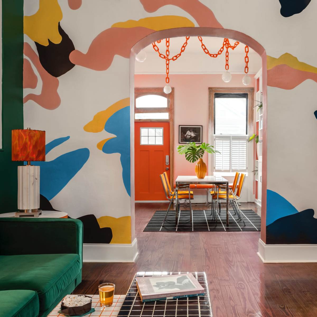 A colorful living room features a green couch, a bold mural on the wall, and an orange door leading to a dining area with a table and chairs.
