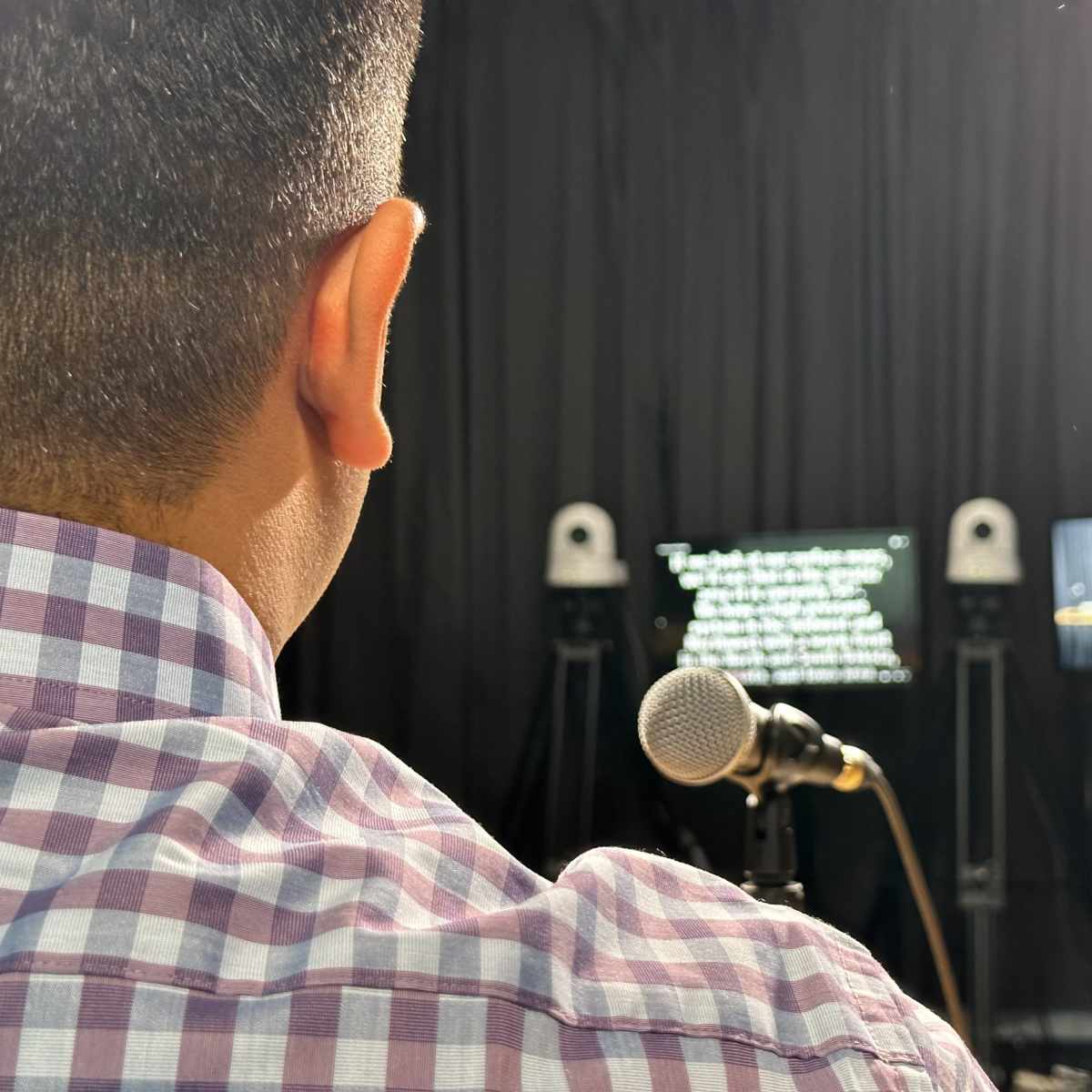 A person in a checkered shirt sits in front of a microphone, looking towards a screen displaying text and another monitor showing a presenter.