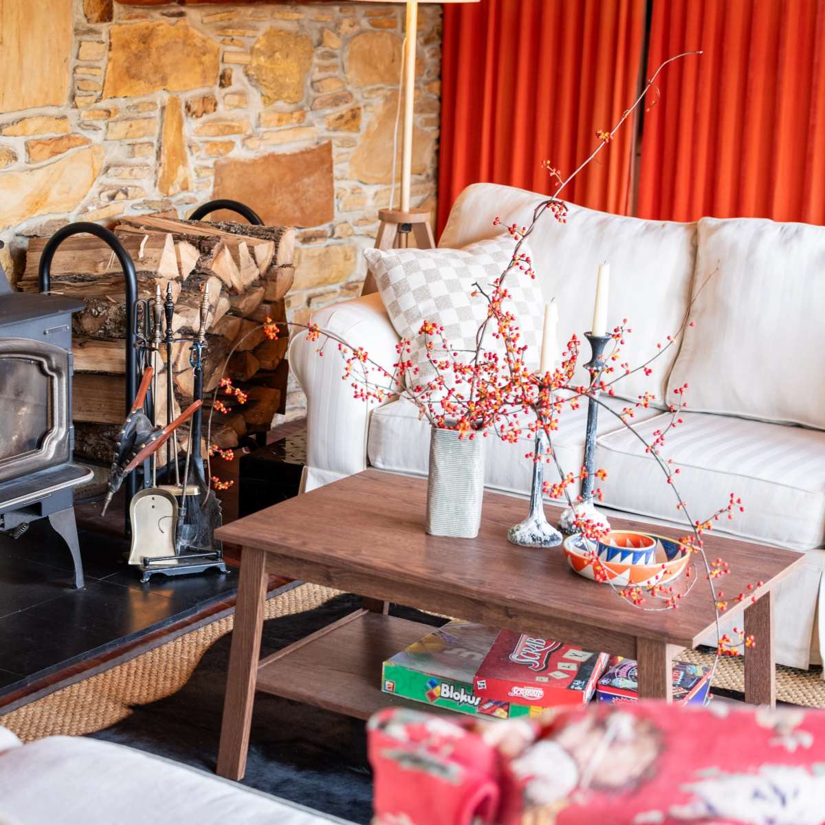 A cozy living room with a black stove, a stone wall, a wooden coffee table, and a sofa adorned with orange curtains in the background.